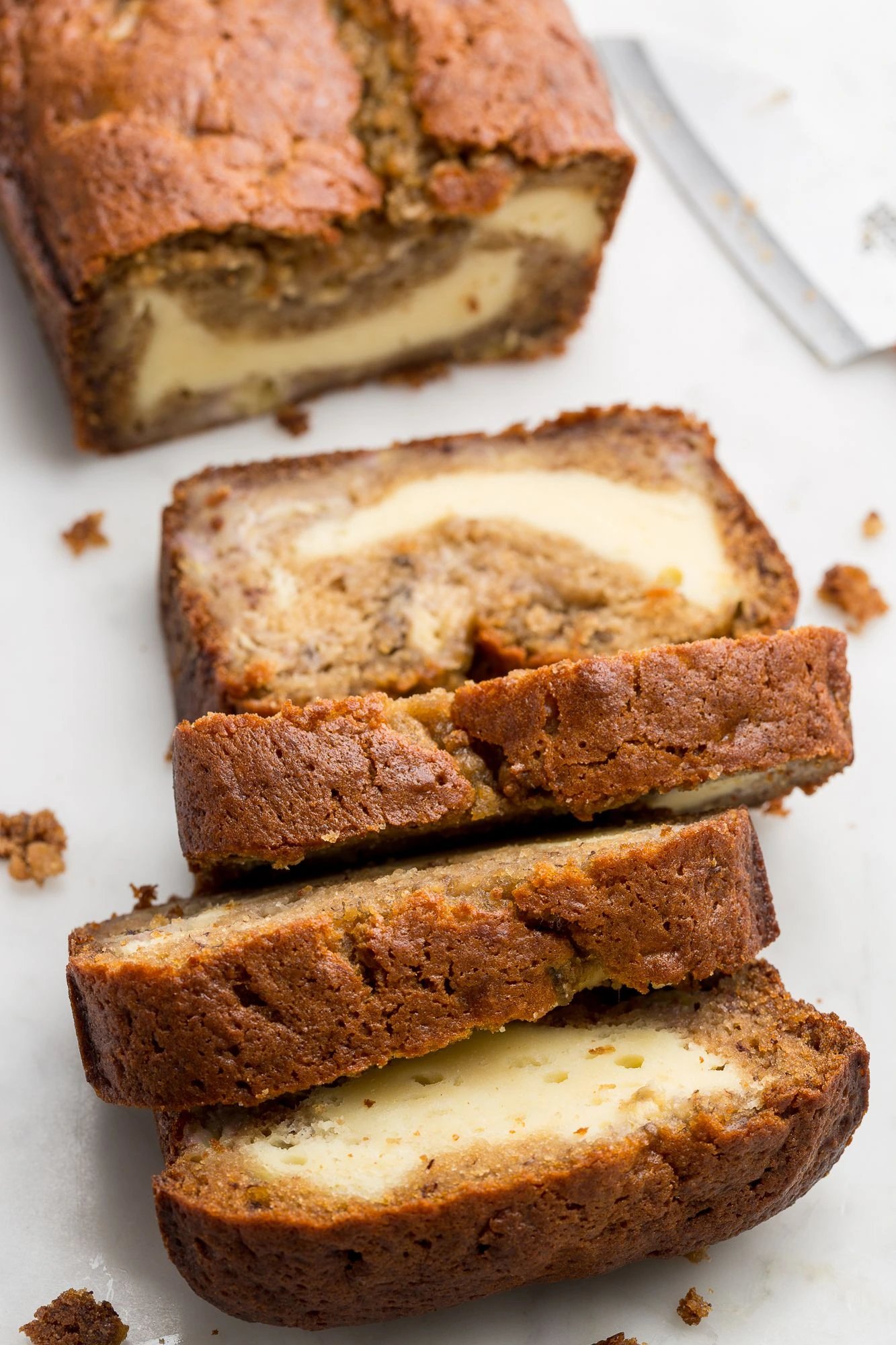 A freshly baked loaf of cheesecake banana bread, sliced to reveal a creamy cheesecake filling swirled into moist banana bread. The golden-brown crust contrasts beautifully with the smooth, pale cheesecake layer.