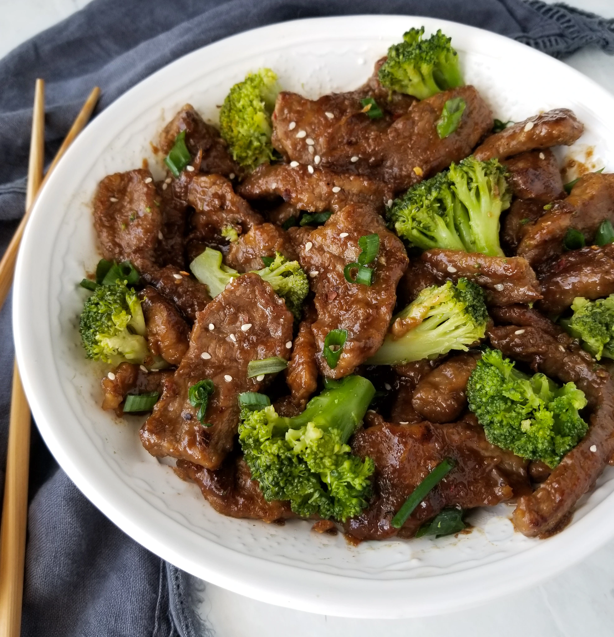 A plate of beef and broccoli stir-fry with tender beef slices, vibrant green broccoli florets, garnished with sesame seeds and green onions on a white plate.