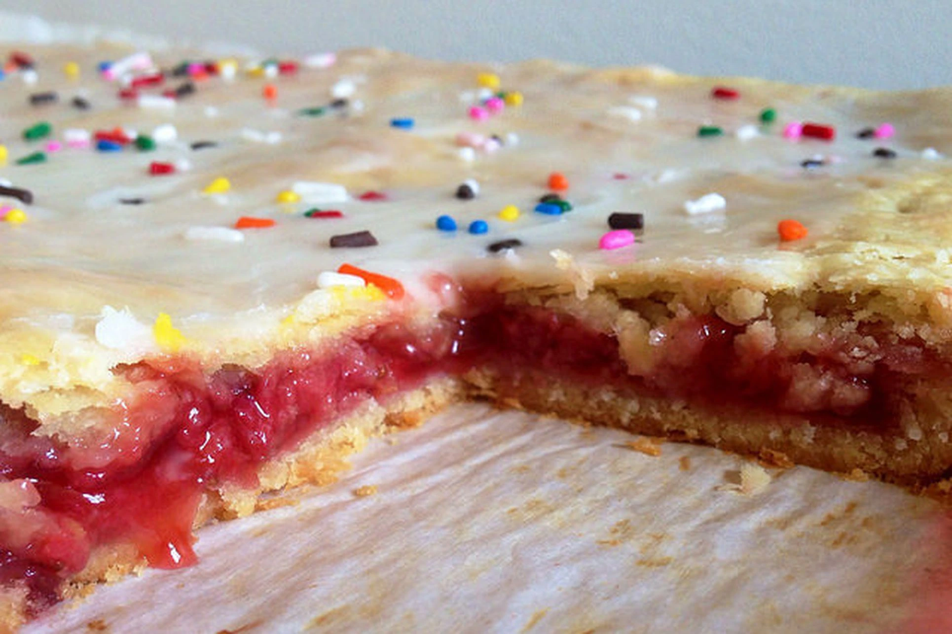 A close-up view of a large rectangular strawberry-filled pastry topped with icing and colorful sprinkles, showing a slice cut out to reveal the filling.