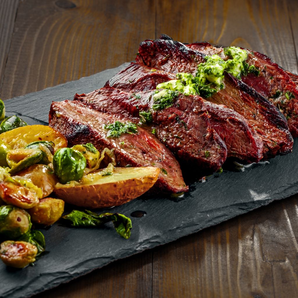 Sliced grilled hanger steak topped with herb butter, served with roasted potatoes and Brussels sprouts on a slate platter.