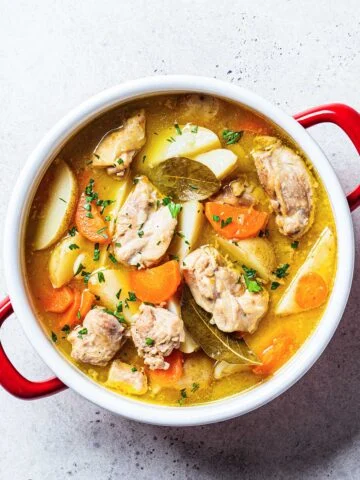 A delicious chicken stew with tender pieces of chicken, carrots, potatoes, and bay leaves in a flavorful golden broth, served in a white pot with red handles.