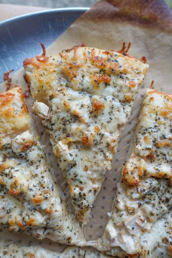 A close-up view of sliced cheesy pizza topped with herbs and chicken on a parchment-lined tray.