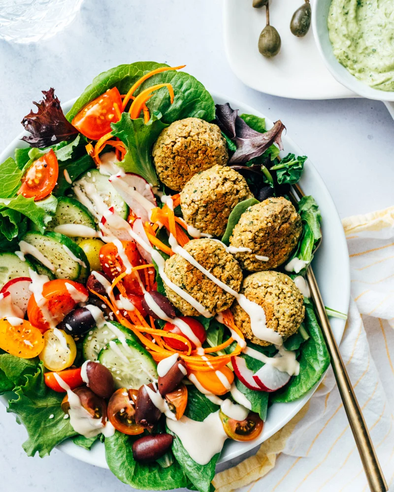 A vibrant falafel salad in a white bowl, featuring crispy falafel balls, fresh greens, cherry tomatoes, cucumbers, shredded carrots, radishes, and Kalamata olives, drizzled with a creamy tahini dressing.