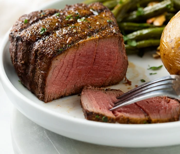 A perfectly cooked filet mignon steak sliced open to reveal a juicy pink interior, served on a white plate with green beans and a baked potato in the background.