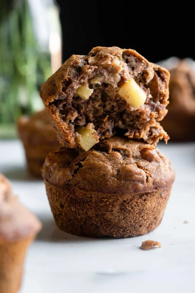 A close-up view of two moist apple banana muffins stacked on a marble surface. The top muffin is broken open, revealing chunks of fresh apple and a soft, spiced crumb.