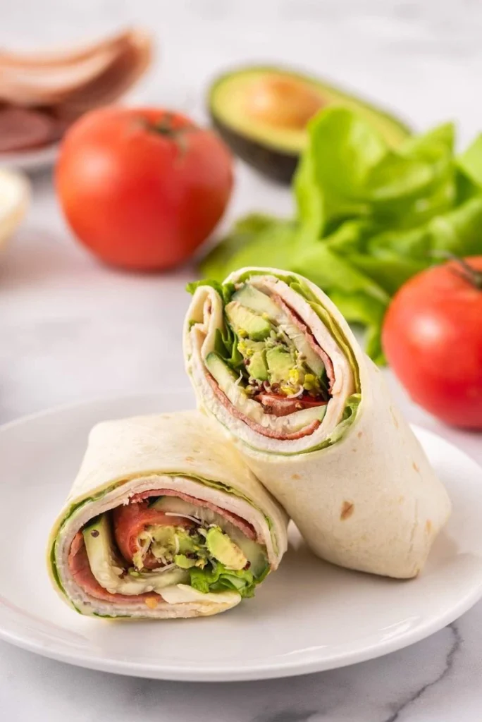 A fresh wrap sliced in half on a white plate, filled with turkey, lettuce, avocado, tomato, and other colorful ingredients, with fresh produce like tomatoes and avocados in the background.