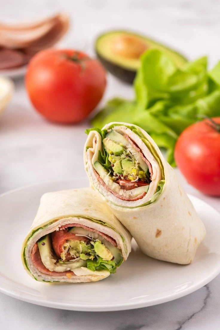 A fresh wrap sliced in half on a white plate, filled with turkey, lettuce, avocado, tomato, and other colorful ingredients, with fresh produce like tomatoes and avocados in the background.