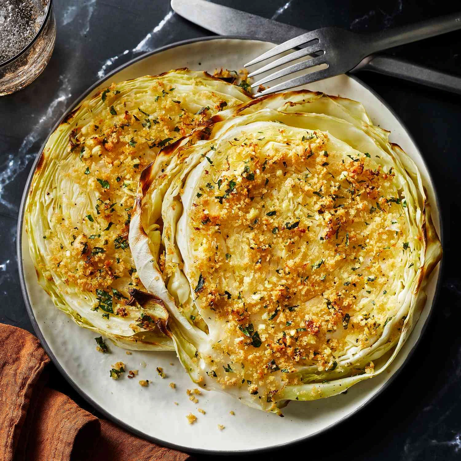 A plate of roasted cabbage steaks topped with golden breadcrumbs, fresh parsley, and a hint of garlic, served with a fork on a dark marble table.