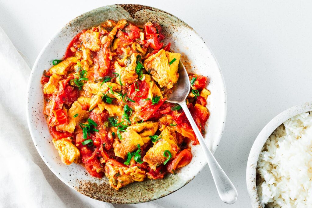 A bowl of Chinese tomato and egg stir-fry garnished with fresh green onions, served alongside a bowl of white rice.