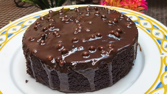 A rich chocolate cake covered with glossy chocolate ganache and decorated with small chocolate pearls, served on a decorative white plate with a colorful border.