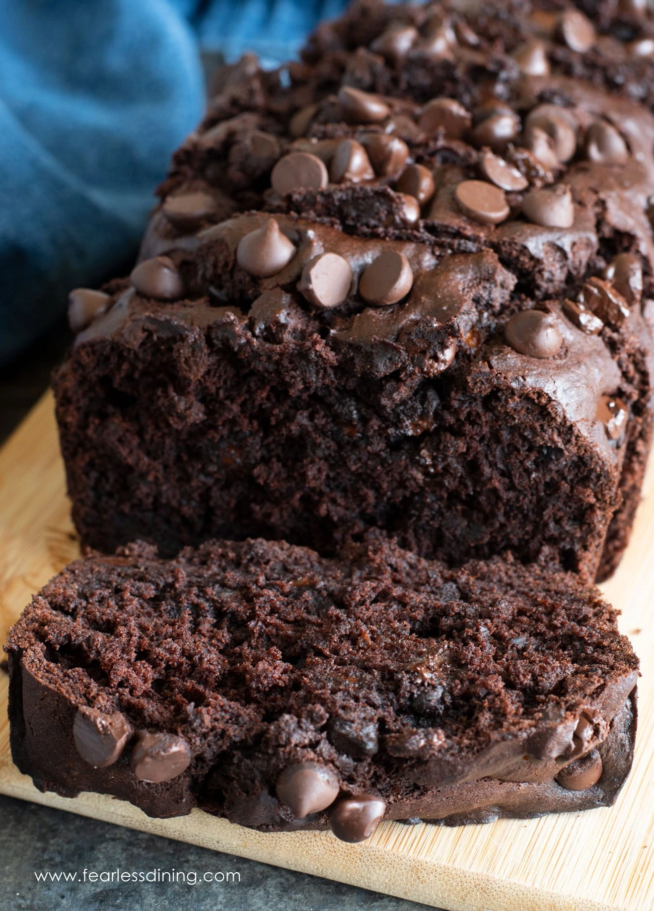 A close-up of a freshly baked chocolate banana bread topped with chocolate chips, placed on a wooden cutting board with one slice partially separated to showcase its moist, rich texture.