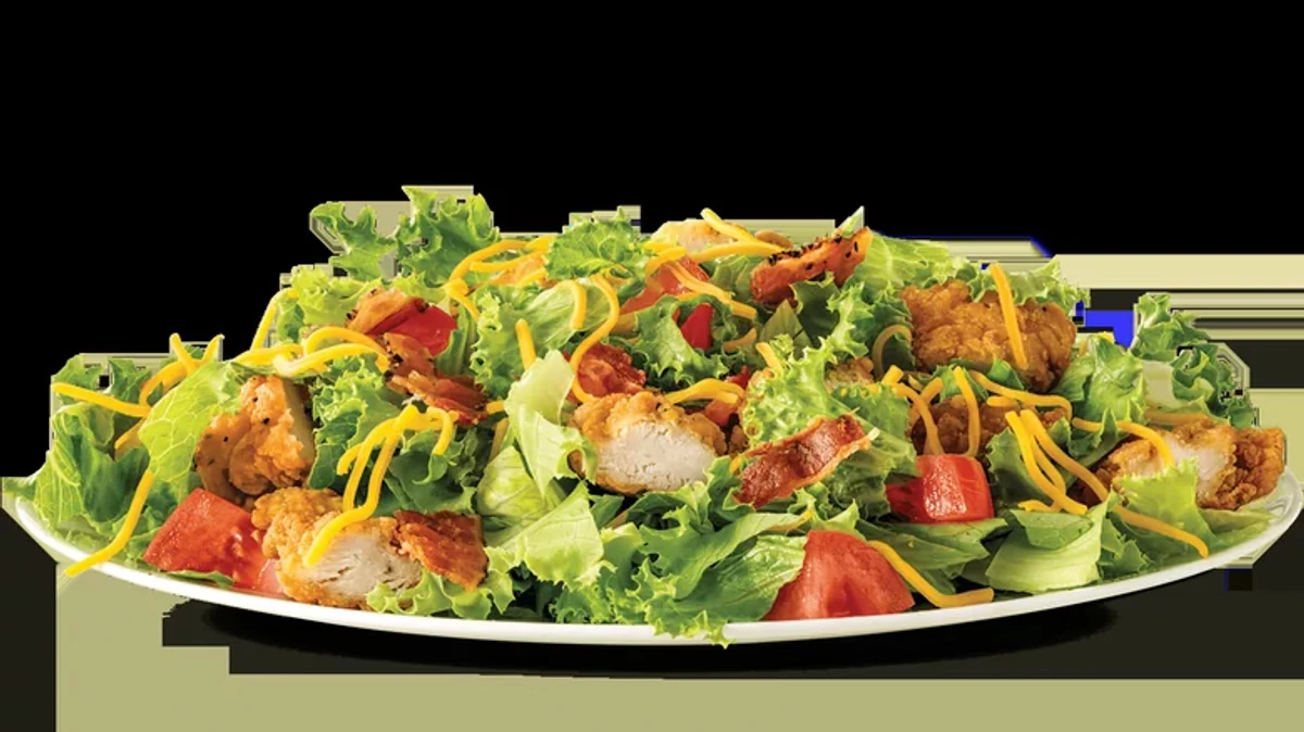 A fresh salad with crispy fried chicken pieces, shredded cheddar cheese, chopped tomatoes, lettuce, and crispy bacon bits, served on a white plate.