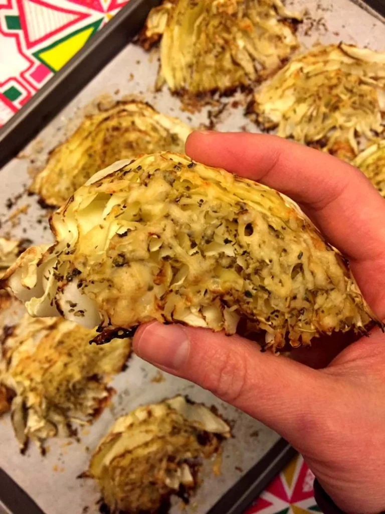 A hand holding a roasted cabbage wedge seasoned with herbs and cheese, with additional roasted cabbage pieces on a tray in the background.