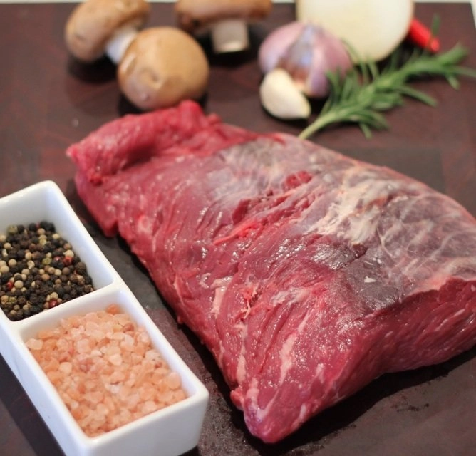 Raw hanger steak on a dark cutting board with ingredients including salt, pepper, mushrooms, garlic, onion, and rosemary.