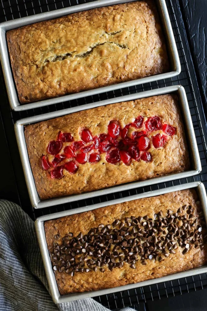 Three freshly baked loaves of banana bread in rectangular pans, placed on a cooling rack. The top loaf is plain, the middle one is topped with slices of fresh strawberries, and the bottom one is topped with chocolate chips.