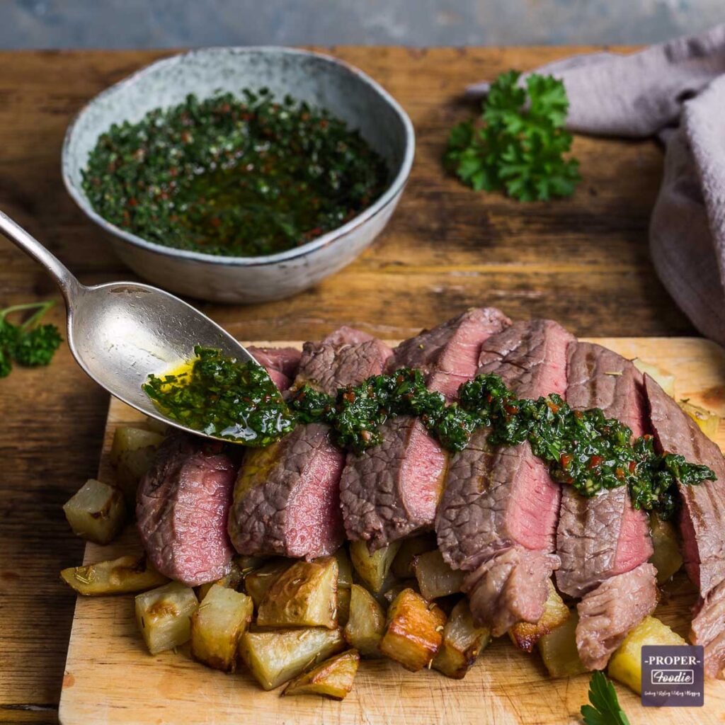 Sliced steak topped with vibrant green chimichurri sauce, served over roasted potatoes on a wooden board, with a bowl of chimichurri in the background.