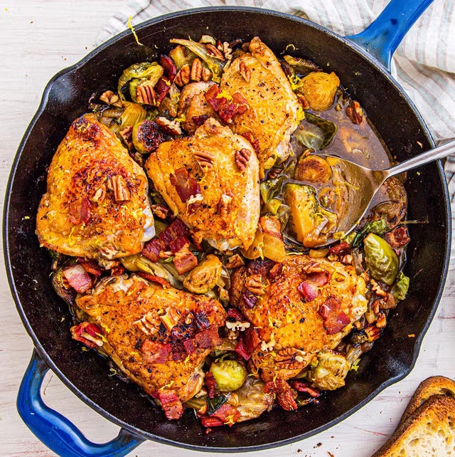 A cast-iron skillet filled with crispy, golden-brown chicken thighs, roasted Brussels sprouts, pecans, bacon bits, and a rich sauce, garnished with seasonings.