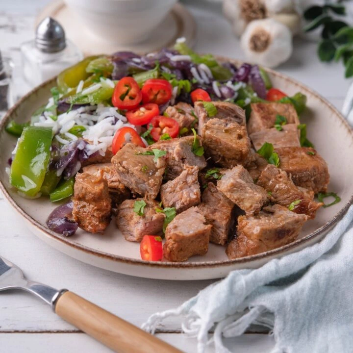 A plate of chipotle steak bites served with white rice, sautéed green bell peppers, red chili slices, and caramelized onions.