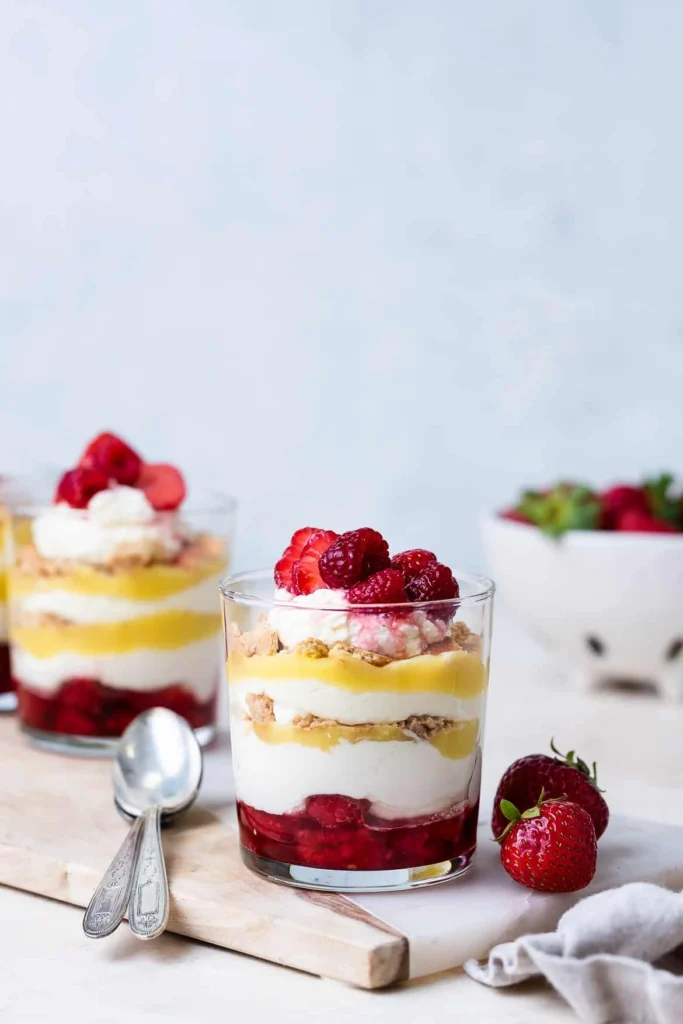 A glass of raspberry lemon parfait layered with creamy yogurt, lemon curd, crushed granola, and fresh raspberries, served on a wooden board with a bowl of berries in the background.