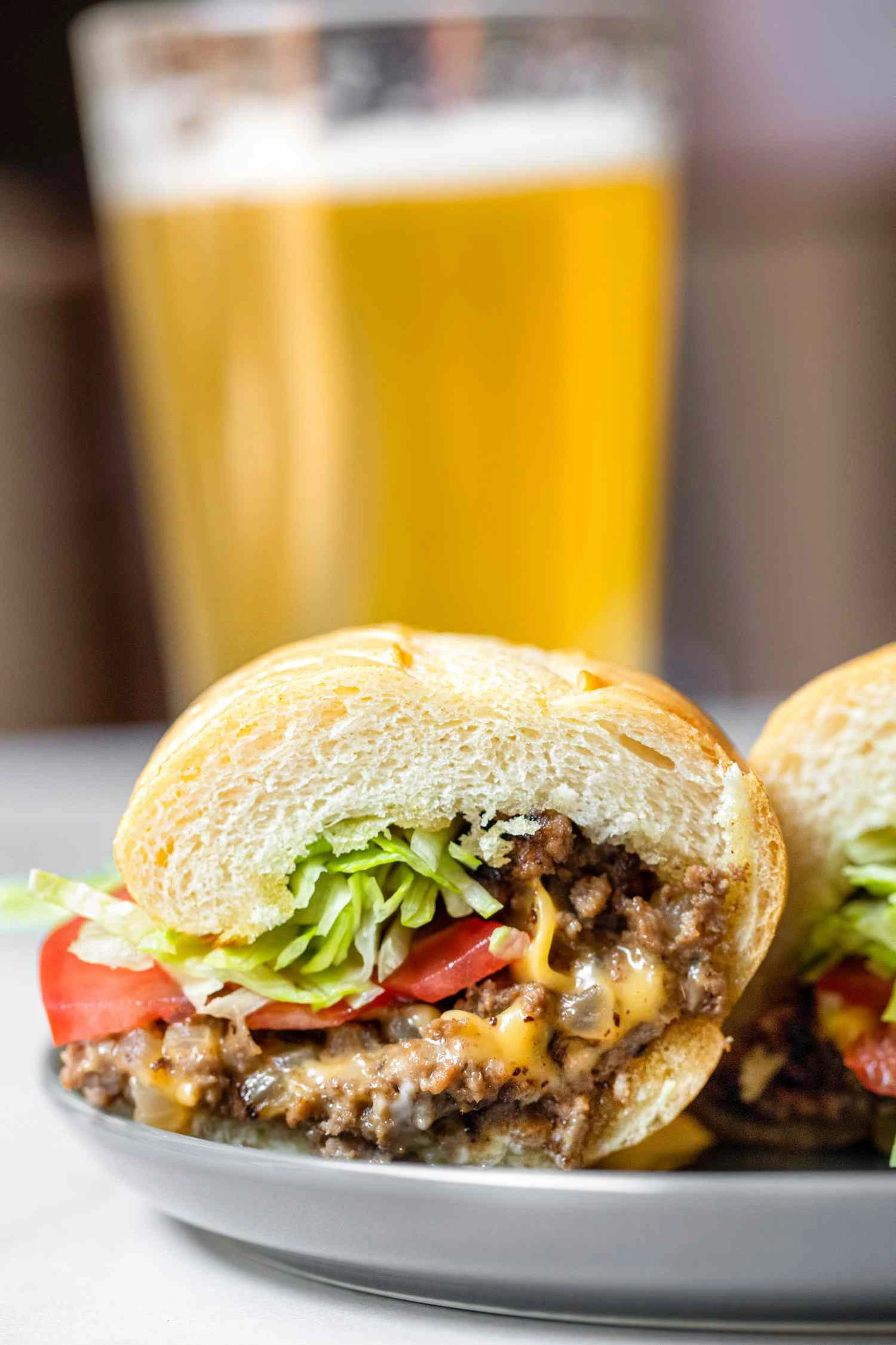 A close-up of a chopped cheese sandwich with melted cheese, seasoned ground beef, lettuce, and tomato in a toasted bun, served on a plate with a glass of beer in the background.