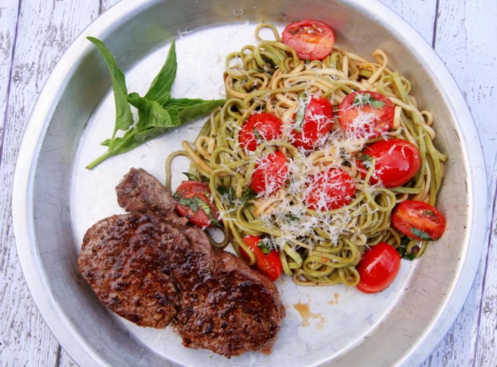 Juicy pan-seared beef steak served with pesto pasta, cherry tomatoes, fresh basil, and grated Parmesan cheese on a silver plate.