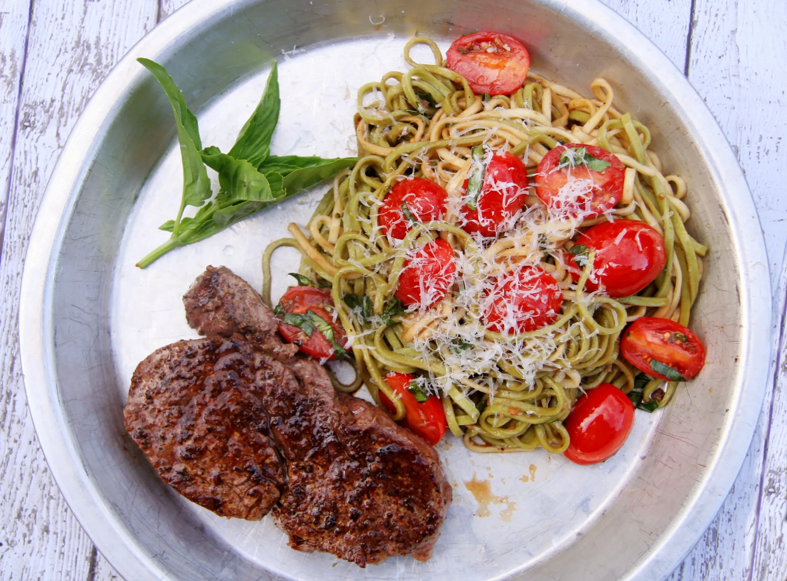 Juicy pan-seared beef steak served with pesto pasta, cherry tomatoes, fresh basil, and grated Parmesan cheese on a silver plate.