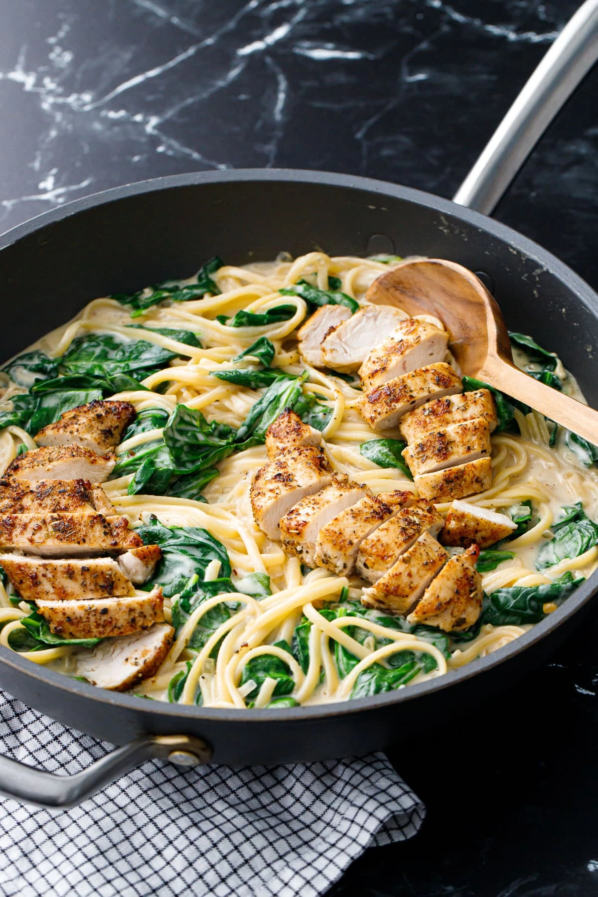 A creamy Chicken Florentine Pasta dish in a black skillet, featuring grilled, sliced chicken breast over spaghetti with fresh spinach in a rich, cheesy sauce. A wooden spoon rests in the pan.