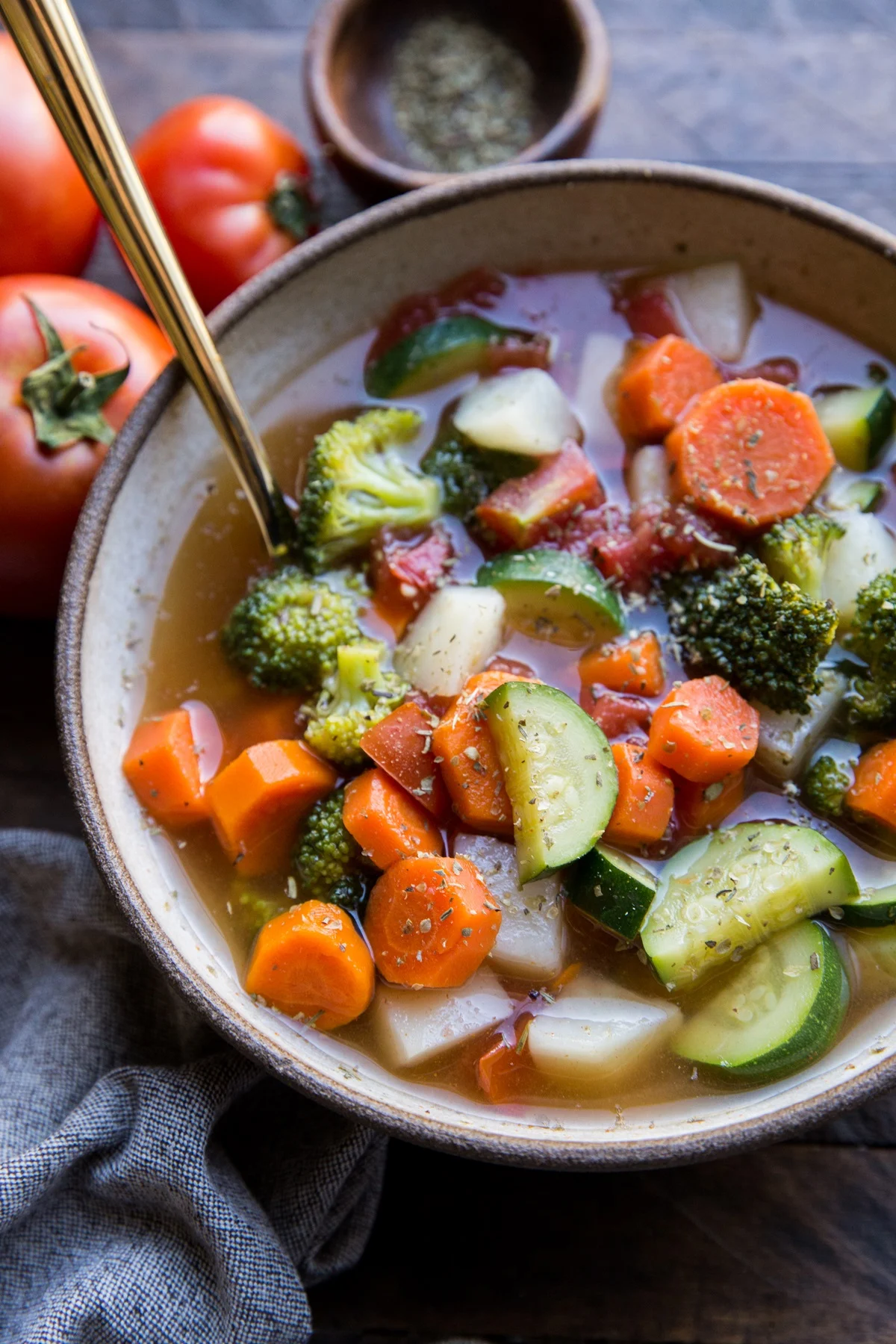 A bowl of homemade vegetable soup with carrots, zucchini, broccoli, and tomatoes in a light broth, garnished with herbs. Fresh tomatoes and seasonings in the background.