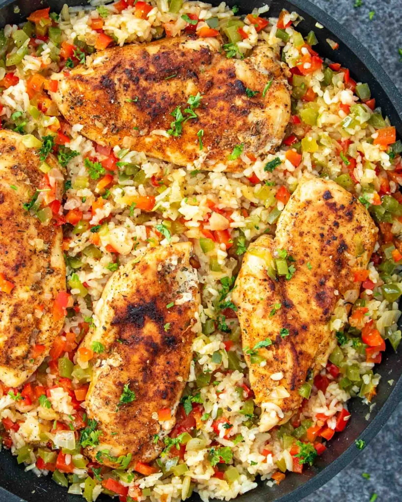 Skillet chicken and rice with golden, seasoned chicken breasts on a bed of fluffy rice mixed with diced red and green bell peppers, garnished with fresh parsley.