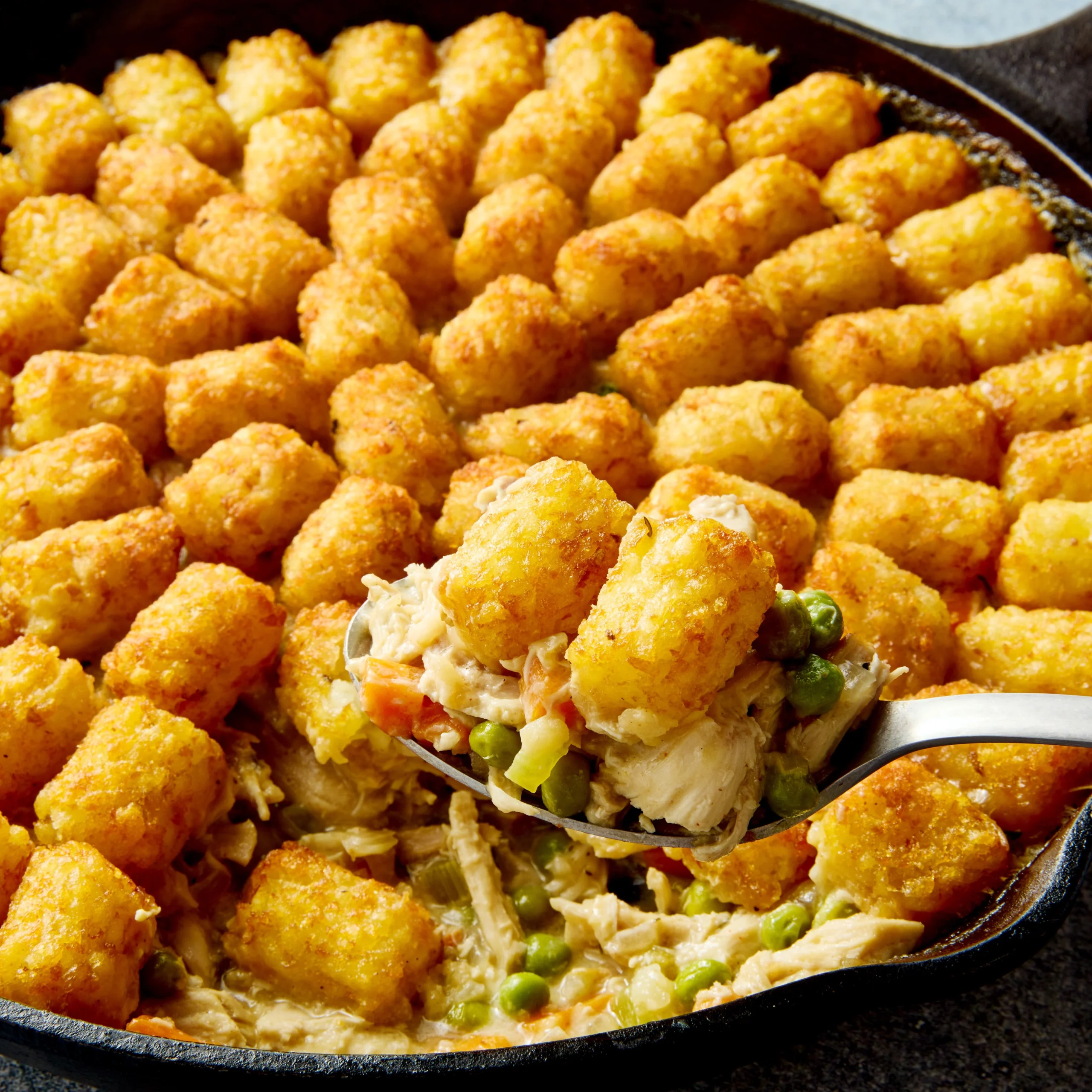 A close-up of a golden, crispy tater tot casserole with a creamy chicken and vegetable filling being scooped from a cast iron skillet.